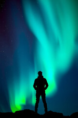 A man in a black jacket stands atop a mountain, his silhouette outlined against the aurora that paints the night sky in mesmerizing hues of blue and green.