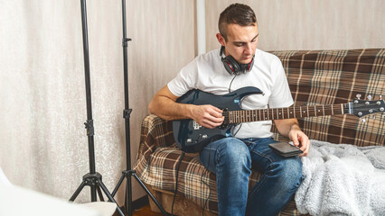 Obraz premium A young man with a smartphone while holding an electric guitar. Musician sitting on a couch and holding guitar