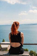 Red-haired woman sitting on a bench, enjoying a seaside view on a sunny day