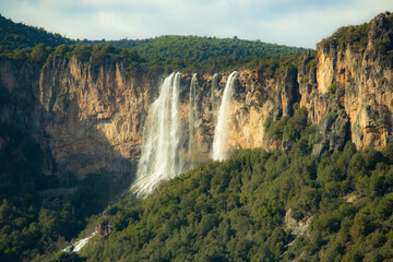 Scenic view of a waterfall flowing down a rocky cliff surrounded by a dense forest, creating a breathtaking natural landscape