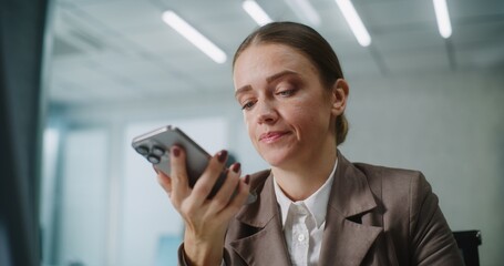 Caucasian woman works in modern office: Female marketing specialist or customer manager sits at workplace, communicates with business partner or client, records voice message on smartphone. E-business