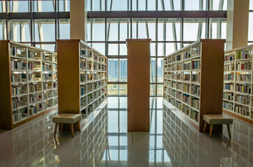 Interior of a modern library featuring symmetrical wooden bookshelves filled with books, and natural light streaming through large floor-to-ceiling windows.A minimalistic and organized study space. © Doublelee