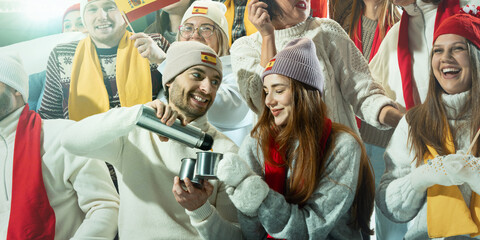 Two sports fans, man and woman sharing warm drink from thermos while enjoying sports event in snowy winter stadium, surrounded by cheerful supporters. Concept of sport, game, competition, emotions