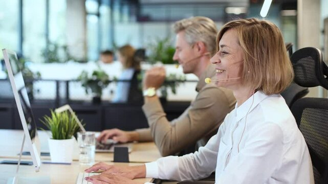 Smiling middle aged female operator call center representative customer support wearing headset working using laptop computer in office