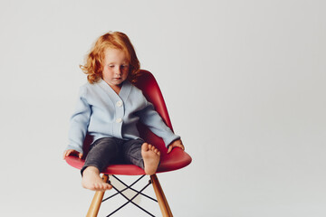 Little red haired girl sitting on a chair in front of a white background innocent beauty in a...