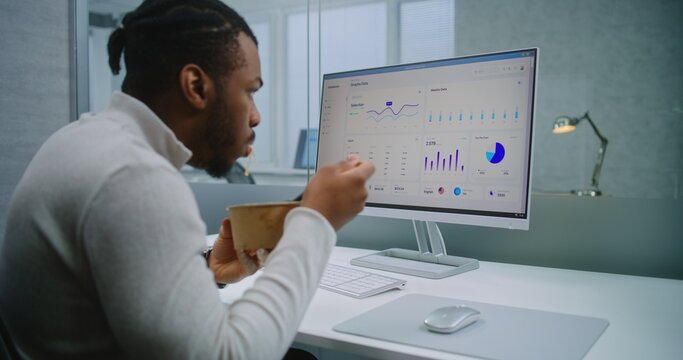 African American man works in modern office of E-commerce company: Businessman or financial specialist eats food from delivery at the workplace, analyzes commercial data and charts using computer.