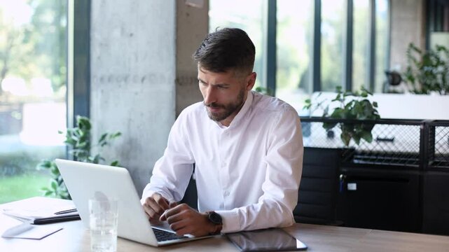 Unhappy frustrated young male holding head by hands sitting with computer behind desk at office.