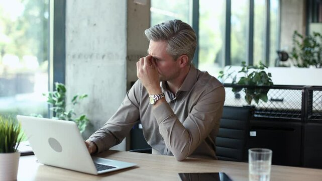 Unhappy frustrated young male holding head by hands sitting with computer behind desk at office.