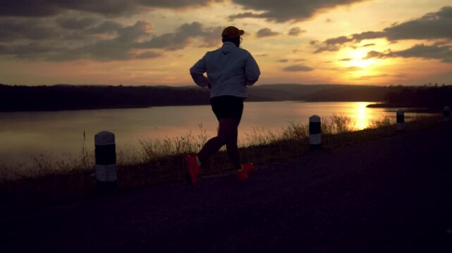 A man runs on a path near a body of water. The sky is dark and the sun is setting