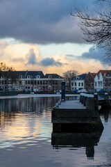 A peaceful canal at sunset with a wooden pier in the foreground, reflecting soft clouds and colorful houses in the calm water, creating a tranquil and scenic atmosphere.