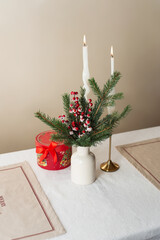 Christmas bouquet with red berries in a ceramic vase, two lit candles and two placemats on a table. The concept of preparation and table setting for the New Year's holiday. Hygge inspired home