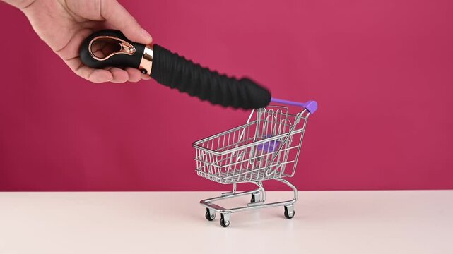 Man putting black sex toy in mini shopping cart on pink background. 