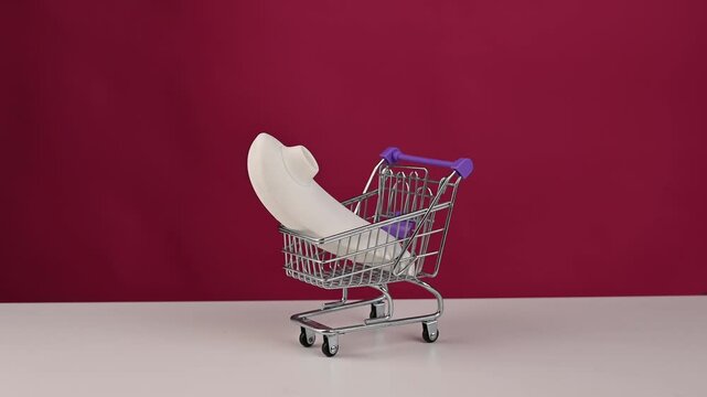 Man putting white vacuum clitoral sex toy into mini shopping cart on pink background. 