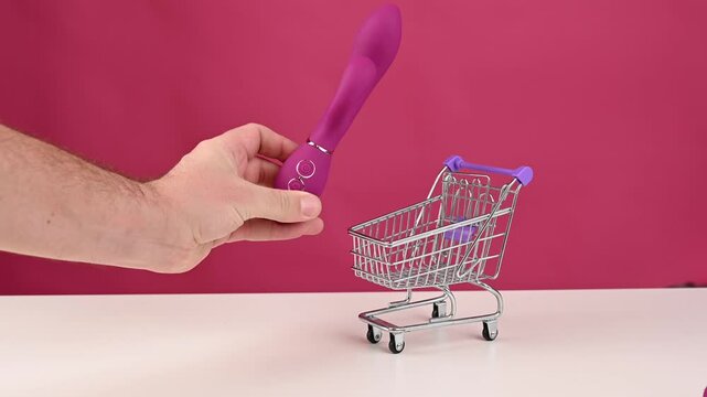 Man putting pink sex toy in mini shopping cart on pink background. 