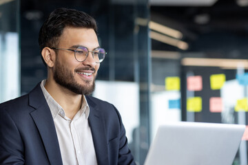 Fototapeta premium A confident businessman is smiling as he works on his laptop in a bright office setting. His expression conveys professionalism and focus in a modern and productive corporate environment.