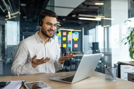 A young businessman is seen participating in a virtual meeting via his laptop, using a headset in a modern office setting. His expressive gestures reflect active communication and professionalism.