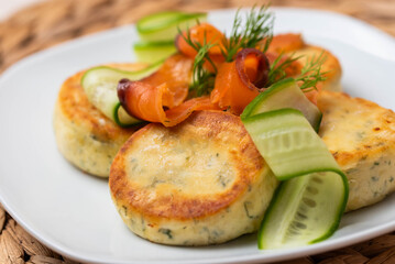 Syrniki with creamy cheese and dill, served on plate with fresh cucumber slices, thinly sliced smoked salmon, and herbs. Concept of traditional home recipes, and balanced meals.