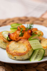Syrniki with creamy cheese and dill, served on plate with fresh cucumber slices, thinly sliced smoked salmon, and herbs. Concept of traditional home recipes, and balanced meals.