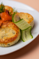 Syrniki with creamy cheese and dill, served on plate with fresh cucumber slices, thinly sliced smoked salmon, and herbs. Concept of traditional home recipes, and balanced meals.