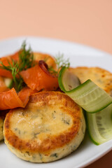 Syrniki with creamy cheese and dill, served on plate with fresh cucumber slices, thinly sliced smoked salmon, and herbs. Concept of traditional home recipes, and balanced meals.
