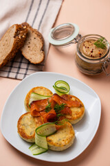 Syrniki (pancakes) with creamy cheese and dill, on plate with fresh cucumber, thinly sliced smoked salmon, and herbs. Jar of homemade pate and fresh bread. Traditional home recipes and gourmet meals.