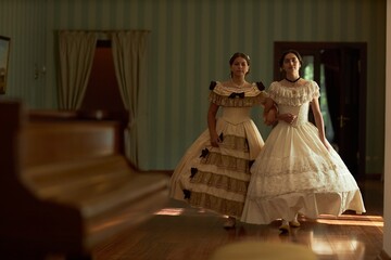 Full length portrait of two young ladies walking together towards camera in voluminous classic dresses across wooden floor in softly lit vintage ballroom, copy space