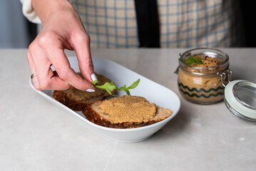 Hands spreading homemade chicken liver pate onto whole-grain bread, topped with fresh tomato slices and arugula. A rustic, savory snack with a touch of elegance and freshness.