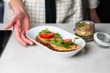 Hands spreading homemade chicken liver pate onto whole-grain bread, topped with fresh tomato slices and arugula. A rustic, savory snack with a touch of elegance and freshness.