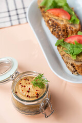 Homemade chicken liver pate in a glass jar, served with whole-grain bread topped with pate, fresh tomato slices, and arugula. A rustic and flavorful dish perfect for breakfast or a light snack.