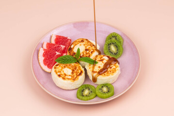 Sweet classic syrniki (cottage cheese pancakes) served on a plate, garnished with slices of grapefruit, fresh kiwi, and a sprig of mint, drizzled with golden sweet syrup. 
