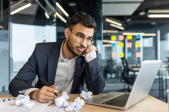 A man businessman in a modern office, looking frustrated at a laptop, surrounded by crumpled paper. He seems to be dealing with a creative block or lack of motivation, seeking inspiration.