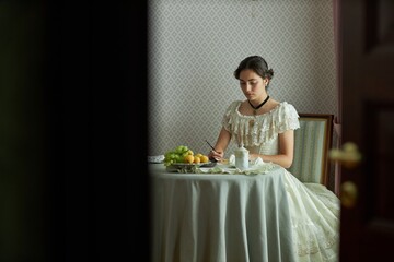 Wide angle view of romantic young lady writing letters at elegant round table in vintage setting...