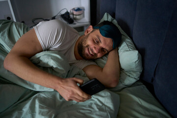  30-year-old man wearing a silk sleep mask is chatting on a smartphone and smiling