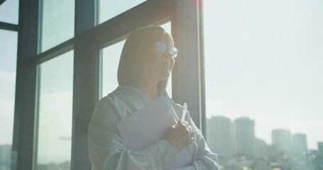Profile of a businesswoman standing by a large office window, holding a clipboard and pen, reflecting on corporate strategy and planning in a sunlit environment
