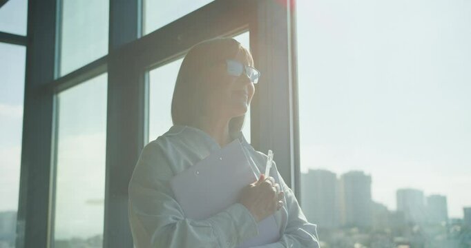 Profile of a businesswoman standing by a large office window, holding a clipboard and pen, reflecting on corporate strategy and planning in a sunlit environment