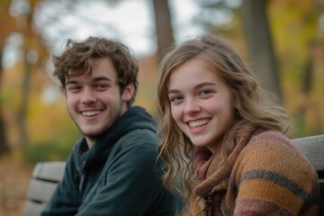 Naklejka premium Two friends smile and laugh while sitting on a bench in a vibrant park during autumn. The background showcases beautiful fall foliage, creating a warm and inviting atmosphere perfect for relaxation an