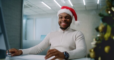 Marketing manager or financial specialist works on computer in modern office. Young successful African American businessman in Santa hat sits at the workplace, smiles and looks at camera. E-commerce.