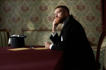 Romantic side view portrait of young gentleman in love looking away pensively sitting at table and writing letter with ink, copy space