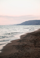 Peaceful beach with gentle waves on a sandy shore, pastel-colored sunset sky, and distant hills creating a tranquil coastal scene, perfect for relaxation and romantic vibes.