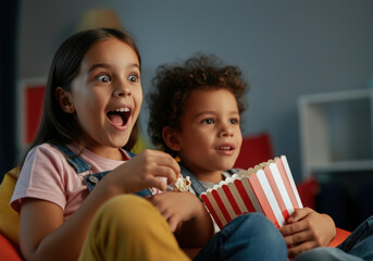 Two young children enjoying a movie night at home, reacting with amazement and eating popcorn