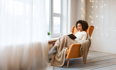 Young woman in warm winter clothes, reading a book in a comfortable armchair by the window at home, covered by a blanket.