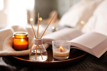 Home diffuser with liquid fragrance and scented candles on tray in bed close up. Cozy aroma atmosphere.