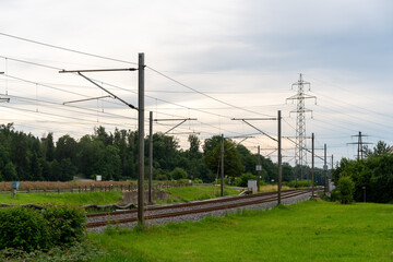 Fototapeta premium Infrastructure of railroad and energy supply come together in this photograph: High voltage pylons of different sizes, and the railroad tracks and overhead contacts. Street besides railroad tracks.