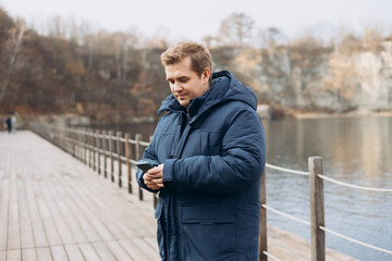 Fototapeta premium Adult 30s man hiker stand outdoors in a natural setting, smile while using his mobile phone outdoors. Happy man standing on the lake wooden pier.