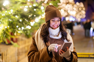 Woman using smartphone on Christmas market, walking and enjoying winter holiday season, blurred lights on background