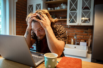 Frustrated young man holding head while studying on laptop at kitchen table