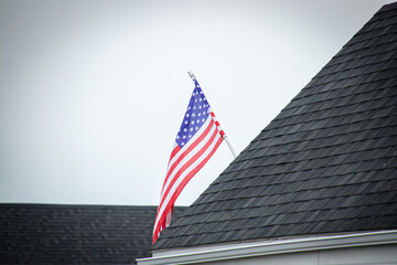 An American flag mounted on a rooftop with dark shingles, set against a cloudy sky, symbolizing patriotism and pride.