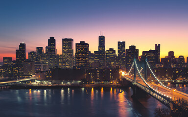 Fototapeta premium City skyline illuminated at dusk with water reflections and bridge in foreground