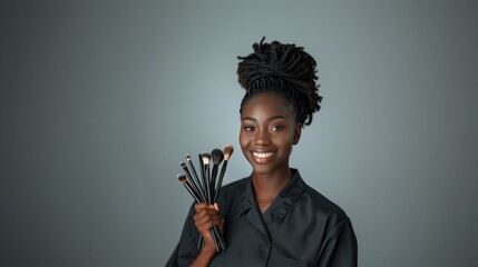 Obraz premium Smiling African American woman makeup artist holding brushes against a neutral gray background, showcasing elegance and professionalism.