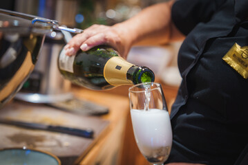 A person in a dark uniform with a gold name tag pours champagne into a flute glass, highlighting luxury and attentive service in a high end environment.